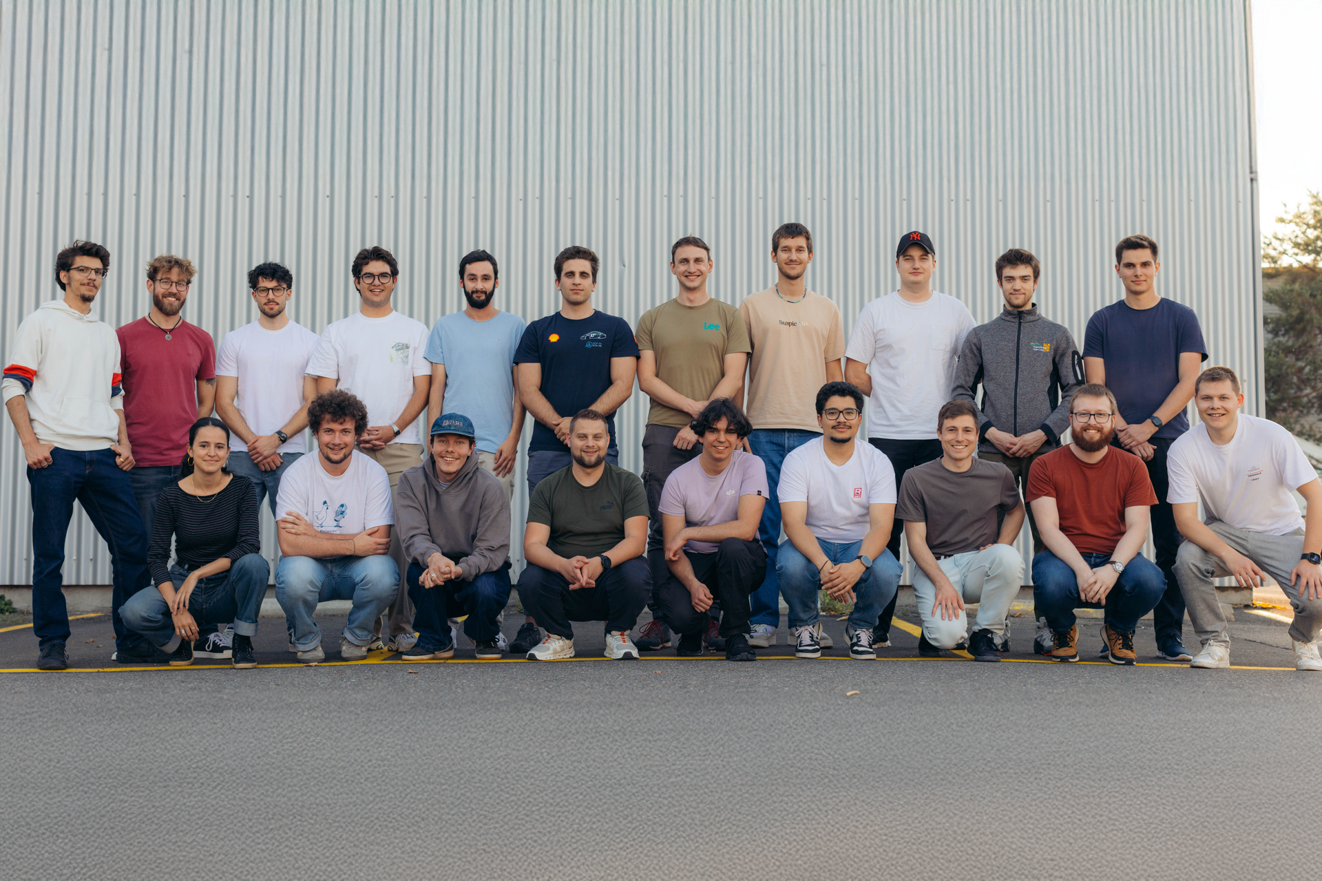Gruppenfoto des Racing-Ost-Teams: Insgesamt 20 Teammitglieder stehen und knien in zwei Reihen vor einer grauen Metallwand. Die Personen tragen überwiegend lässige Kleidung wie T-Shirts, Hoodies und Jeans und lächeln in die Kamera. Das Bild vermittelt ein freundschaftliches, motiviertes Teamgefühl.