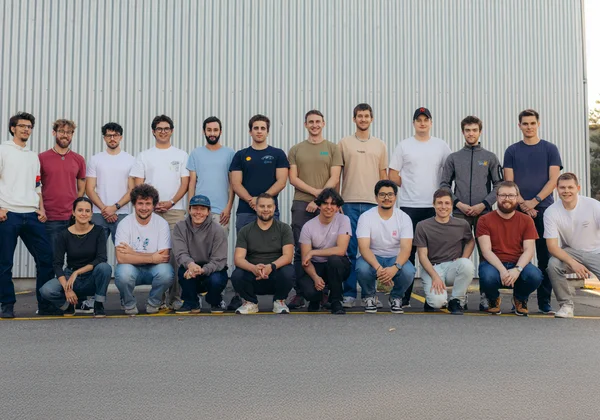 Photo de groupe de l'équipe Racing-Ost : au total, 20 membres de l'équipe sont debout et agenouillés en deux rangées devant un mur métallique gris. La plupart des personnes portent des vêtements décontractés tels que des t-shirts, des sweats à capuche et des jeans, et sourient à la caméra. La photo dégage une impression d'amitié et de motivation au sein de l'équipe.